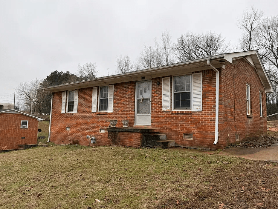 Red brick ranch house with white shutters and a concrete porch under an overcast sky.