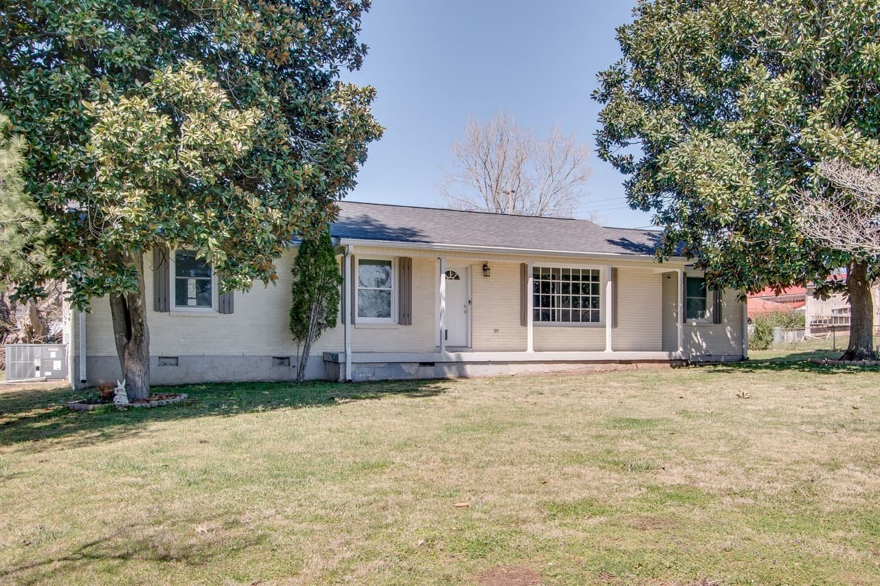 Single-story cream brick ranch home with a front porch, white door, and mature trees.
